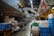 A bustling indoor market scene featuring a variety of crates and containers used for storage and transport. The market is filled with signs written in Japanese, hanging from above, adding to the authenticity and cultural vibe of the setting. The floor is wet, suggesting recent activity or cleaning. Workers are seen in the midground, busy with their tasks in this organized yet crowded space.