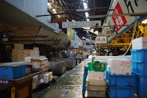 A bustling indoor market scene featuring a variety of crates and containers used for storage and transport. The market is filled with signs written in Japanese, hanging from above, adding to the authenticity and cultural vibe of the setting. The floor is wet, suggesting recent activity or cleaning. Workers are seen in the midground, busy with their tasks in this organized yet crowded space.