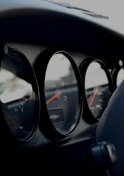 A close-up view of the dashboard in a car, focusing on the analog dials. The dials have black backgrounds with orange and white indicators, showing various measurements. The lighting suggests a dimly lit or shadowed environment, highlighting the curvature and material texture of the dashboard.