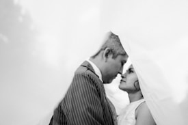 A black and white photograph features a couple standing very close to each other with their foreheads touching, creating a sense of intimacy. The scene is framed by soft, out-of-focus fabric or veil-like material, adding an ethereal quality. The man is wearing a pinstripe suit jacket and the woman has her hair styled elegantly.