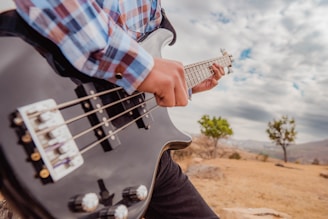 A person playing a black electric bass guitar outdoors, wearing a plaid shirt. The background features an open landscape with dry grass, scattered stones, and a few green trees under a cloudy sky.