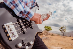 A person playing a black electric bass guitar outdoors, wearing a plaid shirt. The background features an open landscape with dry grass, scattered stones, and a few green trees under a cloudy sky.