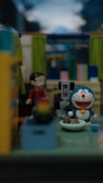 A small room with colorful furnishings and a variety of objects. In the center, a blue and white toy cat with a round smiling face is sitting on the floor. Nearby, a small human-like figure dressed in red is standing. The background shows brightly colored curtains and shelves with various items, creating a vibrant and playful atmosphere.