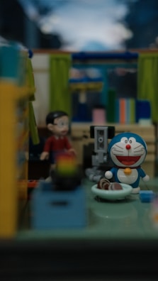 A small room with colorful furnishings and a variety of objects. In the center, a blue and white toy cat with a round smiling face is sitting on the floor. Nearby, a small human-like figure dressed in red is standing. The background shows brightly colored curtains and shelves with various items, creating a vibrant and playful atmosphere.