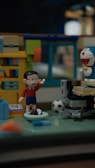 A diorama features toy figures in a miniature room setting. One figure raises its hand, wearing a red shirt and blue shorts. Nearby, a robot-like figure with a blue and white design and a red nose is positioned. The room includes a bookshelf with colorful books, a computer desk, and a soccer ball on the floor.