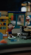 A diorama features toy figures in a miniature room setting. One figure raises its hand, wearing a red shirt and blue shorts. Nearby, a robot-like figure with a blue and white design and a red nose is positioned. The room includes a bookshelf with colorful books, a computer desk, and a soccer ball on the floor.