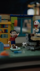 A diorama features toy figures in a miniature room setting. One figure raises its hand, wearing a red shirt and blue shorts. Nearby, a robot-like figure with a blue and white design and a red nose is positioned. The room includes a bookshelf with colorful books, a computer desk, and a soccer ball on the floor.