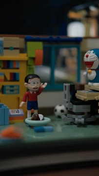 A diorama features toy figures in a miniature room setting. One figure raises its hand, wearing a red shirt and blue shorts. Nearby, a robot-like figure with a blue and white design and a red nose is positioned. The room includes a bookshelf with colorful books, a computer desk, and a soccer ball on the floor.