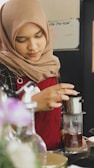 A person wearing a tan headscarf and red apron is preparing a drink using a drip coffee maker. Their focus is on the preparation, suggesting a careful and precise process. The setting appears to be a cafe or kitchen.
