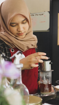 A person wearing a tan headscarf and red apron is preparing a drink using a drip coffee maker. Their focus is on the preparation, suggesting a careful and precise process. The setting appears to be a cafe or kitchen.