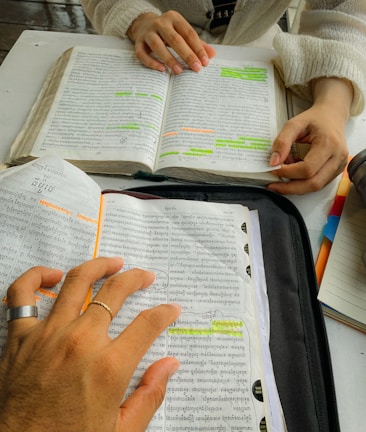 Two people are studying with books open on a table, one person has highlighted sections in orange and green. Their hands are visible, and there are notebooks and pens nearby.