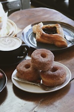 A plate with three stacked sugar-coated doughnuts is placed on a wooden table, accompanied by a fork. Nearby, there is a cappuccino in a black mug with a frothy top, and a pastry featuring a dark fruit filling on a blue plate.