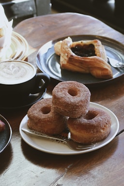A plate with three stacked sugar-coated doughnuts is placed on a wooden table, accompanied by a fork. Nearby, there is a cappuccino in a black mug with a frothy top, and a pastry featuring a dark fruit filling on a blue plate.