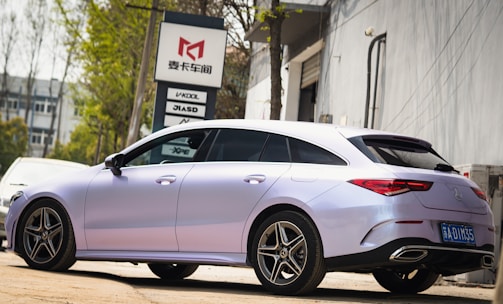 A sleek, modern car with a glossy metallic finish is parked outdoors, adjacent to a building. The vehicle has a compact, aerodynamic design with stylish alloy wheels. In the background, a sign with various brand logos is visible, along with some foliage and another parked car.