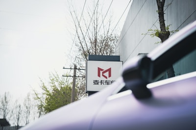 A sign with Chinese characters and a stylized red logo on a white background is prominently displayed. In the foreground, a part of a car is visible. Leafless trees and power lines are present in the background. There is a modern building with minimal architectural features visible next to the sign.