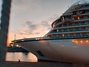 A large cruise ship named Viking Mars is docked at a port during sunset. The ship's lights are on, illuminating its multiple decks and balconies. Calm water reflects the orange and blue hues of the sky as some clouds drift above.