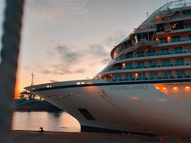 A large cruise ship named Viking Mars is docked at a port during sunset. The ship's lights are on, illuminating its multiple decks and balconies. Calm water reflects the orange and blue hues of the sky as some clouds drift above.