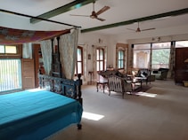 A spacious, well-lit room with large windows and elegant wooden furniture. There is a blue-covered canopy bed in the foreground, surrounded by rich wood detailing. In the background, a sitting area features a set of striped upholstered chairs and a small table. The room has a vintage style, with light pouring in through windows dressed with curtains. Ceilings have green beams and are equipped with ceiling fans. The floor is a light neutral color, creating a warm ambiance.