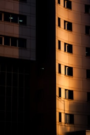 The image features a section of a tall building with a curved facade bathed in warm, golden sunlight. A significant contrast between light and shadow is evident, highlighting the architectural details and creating a dramatic effect. The building's windows are prominently displayed, with some reflecting light while others remain dark.
