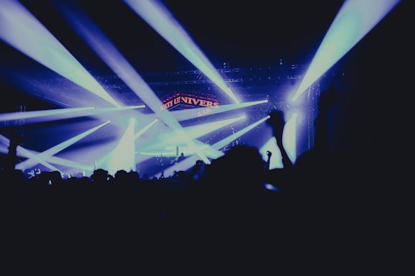 A lively concert scene with bright stage lights illuminating the area. The silhouette of an enthusiastic crowd with raised hands can be seen in the foreground, creating a dynamic and energetic atmosphere.