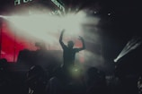 A person is silhouetted against bright stage lights at a concert or music event. The atmosphere is filled with fog or smoke, enhancing the vivid lighting effects. The scene is lively, with a crowd surrounding the main subject. The background features a DJ booth and vibrant lighting, suggesting a dynamic performance.