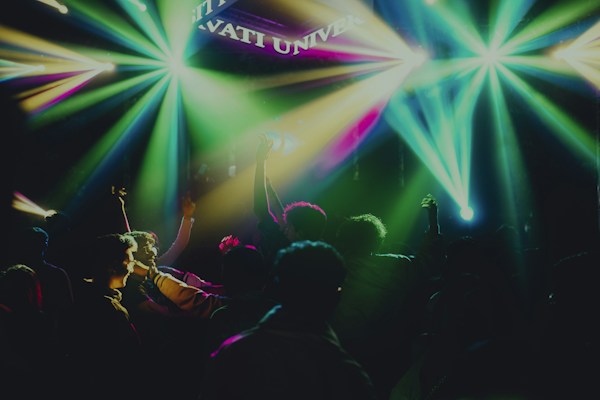 A vibrant and energetic crowd gathered in a dimly lit venue, with vibrant multicolored lights beaming across the space. People appear to be dancing and enjoying a lively atmosphere, with hands raised and faces illuminated by the colorful lighting.