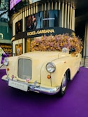 A vintage cream-colored car is adorned with a large bouquet of dried flowers on its roof. The car is positioned on a purple carpet in front of a Dolce & Gabbana store, visible in the background. The license plate reads 'Jo Malone London'. The setting gives a luxurious and artistic display, likely for promotional purposes.