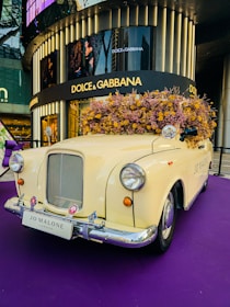 A vintage cream-colored car is adorned with a large bouquet of dried flowers on its roof. The car is positioned on a purple carpet in front of a Dolce & Gabbana store, visible in the background. The license plate reads 'Jo Malone London'. The setting gives a luxurious and artistic display, likely for promotional purposes.