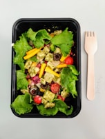 A fresh salad presented in a black rectangular container, featuring vibrant green lettuce leaves, cherry tomatoes, sliced black olives, yellow bell pepper strips, and cubes of pesto-marinated cheese or tofu. A wooden fork is placed to the right of the container on a light surface.