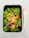 A fresh salad presented in a black rectangular container, featuring vibrant green lettuce leaves, cherry tomatoes, sliced black olives, yellow bell pepper strips, and cubes of pesto-marinated cheese or tofu. A wooden fork is placed to the right of the container on a light surface.