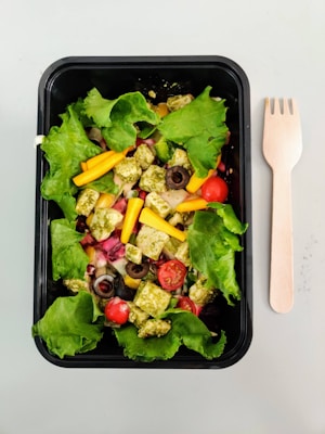 A fresh salad presented in a black rectangular container, featuring vibrant green lettuce leaves, cherry tomatoes, sliced black olives, yellow bell pepper strips, and cubes of pesto-marinated cheese or tofu. A wooden fork is placed to the right of the container on a light surface.