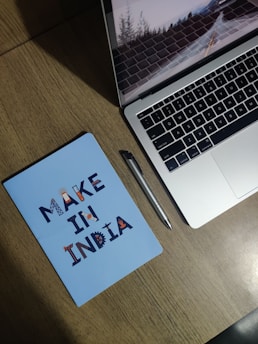A wooden desk with a silver laptop partially open, showing a screen saver of a road with trees. Next to it is a silver pen and a blue notebook with the text 'MAKE IN INDIA' in decorative letters.