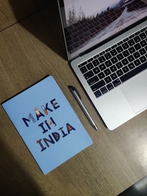 A wooden desk with a silver laptop partially open, showing a screen saver of a road with trees. Next to it is a silver pen and a blue notebook with the text 'MAKE IN INDIA' in decorative letters.