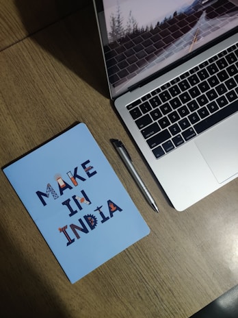 A wooden desk with a silver laptop partially open, showing a screen saver of a road with trees. Next to it is a silver pen and a blue notebook with the text 'MAKE IN INDIA' in decorative letters.