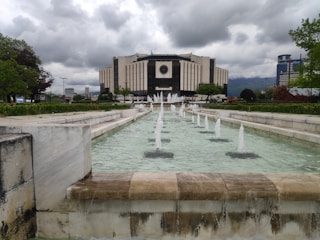 A modern building with a large circular emblem is situated behind a series of cascading fountains. The building is surrounded by greenery and trees. The sky is overcast with dark clouds, creating a dramatic atmosphere.