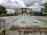 A modern building with a large circular emblem is situated behind a series of cascading fountains. The building is surrounded by greenery and trees. The sky is overcast with dark clouds, creating a dramatic atmosphere.