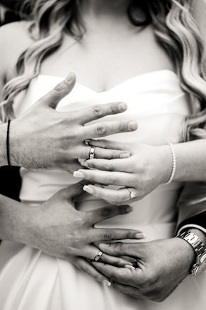 Multiple hands with rings are gently placed around a bride in a wedding dress, capturing an intimate moment. The image is in black and white, focusing on the close-up of hands and rings, symbolizing marriage and connection.