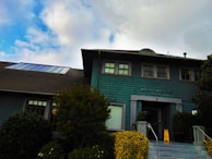 A two-story building with teal-colored shingles and a dark roof, featuring several windows and a main entrance. A sign on the front reads 'BERKELEY TENNIS CLUB EST. 1906'. The entrance is accessed by steps lined with shrubs and a caution sign is placed near the doorway. The background shows a partly cloudy sky.