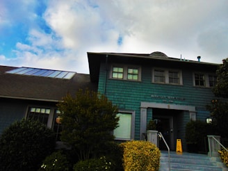 A two-story building with teal-colored shingles and a dark roof, featuring several windows and a main entrance. A sign on the front reads 'BERKELEY TENNIS CLUB EST. 1906'. The entrance is accessed by steps lined with shrubs and a caution sign is placed near the doorway. The background shows a partly cloudy sky.