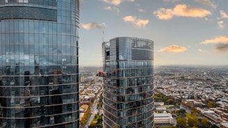 Aerial view of two modern high-rise glass buildings with a window cleaning platform suspended between them. The cityscape stretches out below with numerous smaller buildings, streets, and patches of greenery visible. The sky is mostly clear with a few scattered clouds reflecting off the glass surfaces.