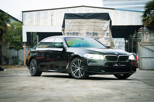 A sleek black luxury sedan is parked in a somewhat industrial setting. The background includes a partially open warehouse with a tarp featuring some text, and there are trees and foliage visible on the sides. The car features prominent headlights and clean, shiny bodywork.