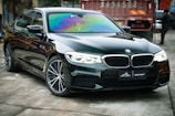 A sleek black sedan with shiny, reflective surfaces is parked indoors. The windshield displays a range of iridescent colors, suggesting a tint or coating. In the background, a red industrial container with numbers and caution stripes adds an urban or industrial feel to the scene.