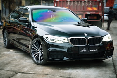 A sleek black sedan with shiny, reflective surfaces is parked indoors. The windshield displays a range of iridescent colors, suggesting a tint or coating. In the background, a red industrial container with numbers and caution stripes adds an urban or industrial feel to the scene.