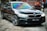 A sleek black sedan with shiny, reflective surfaces is parked indoors. The windshield displays a range of iridescent colors, suggesting a tint or coating. In the background, a red industrial container with numbers and caution stripes adds an urban or industrial feel to the scene.