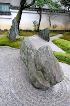 Large textured stones are artistically arranged on raked gravel in a serene Zen garden, surrounded by lush green moss. Trees with twisted trunks and sparse leaves stand amidst the garden, enclosed by a traditional wall.