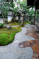 A serene Japanese garden featuring a winding gravel path bordered by lush green moss and stone tiles. Mature trees with intricate branches provide shade, while a traditional stone lantern is visible among the foliage. Natural rocks are scattered throughout the landscape, contributing to the tranquil atmosphere.