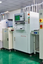 A laboratory setting with several advanced machines, including a prominent white machine labeled 'SINIC-TEK' featuring a large monitor on top displaying technical graphics. Beside it, another machine displays the label 'GWEI' with a smaller screen. The setting has a high-tech and industrial appearance with a polished green floor and metallic curtains.