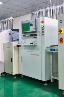 A laboratory setting with several advanced machines, including a prominent white machine labeled 'SINIC-TEK' featuring a large monitor on top displaying technical graphics. Beside it, another machine displays the label 'GWEI' with a smaller screen. The setting has a high-tech and industrial appearance with a polished green floor and metallic curtains.