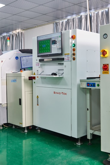 A laboratory setting with several advanced machines, including a prominent white machine labeled 'SINIC-TEK' featuring a large monitor on top displaying technical graphics. Beside it, another machine displays the label 'GWEI' with a smaller screen. The setting has a high-tech and industrial appearance with a polished green floor and metallic curtains.