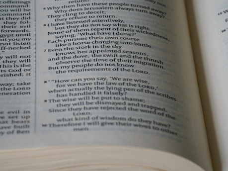 A close-up view of an open book displaying text with religious or biblical content. The text is printed in two columns and includes verse numbers. The pages are slightly curved, indicating the book is being held open.